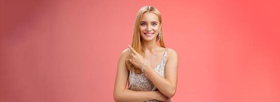 Confident attractive feminine glamour blond young woman in silver glittering dress smiling broadly pointing up discuss recent news promo sales standing friendly grinning camera red background photo