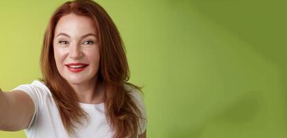 Close-up joyful enthusiastic redhead alluring middle-aged woman extand arm towards camera taking selfie smartphone smiling broadly posing photograph taking picture device green background photo