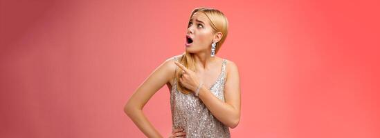 Shocked displeased bothered arrogant blond glamour woman in silver glittering dress turning upper right corner pointing complaining strange noise come upstairs, standing questioned red background photo