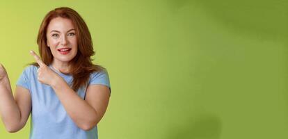 Curious enthusiastic gorgeous redhead female intrigued pointing upper left corner asking question interesting product talk shop assistant consulting promo stand green background blue t-shirt photo