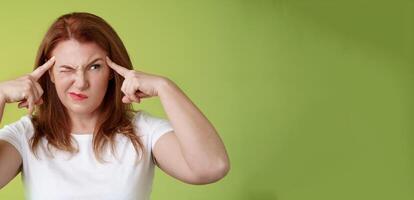 Difficult decision. Perplexed hesitant redhead middle-aged 50s woman trying solve puzzle look puzzled troubled smirk close eyes peek right touch temples thoughtful thinking intense green background photo