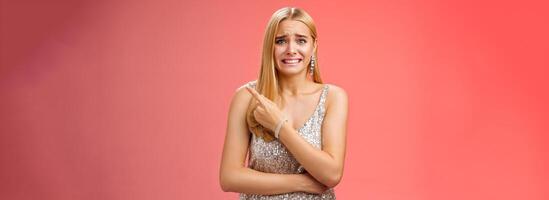 Nervous hesitant attractive worried blond woman cringing frowning clench teeth pointing behind seek friends help see ex-boyfriend unwilling be seen, standing anxious awkward party red background photo