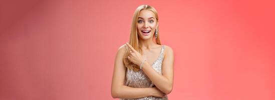Joyful surprised good-looking blond woman talking friend during party smiling broadly look camera amused wondered pointing upper right corner curious discuss awesome room design, red background photo