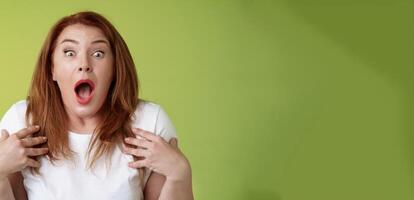 Shocked panicking redhead middle-aged woman gasping drop jaw open mouth stare camera freak-out anxious pointing herself impressed terrified frustrated nervously react green background photo