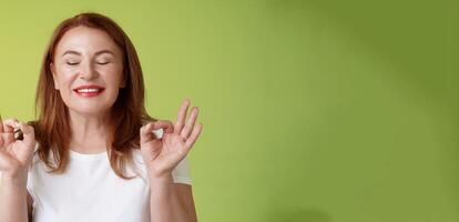 Close to perfection. Close-up peaceful relaxed redhead happy woman closed eyes pure delighted smile show zen peace satisfaction gesture meditating reach nirvana calm stand green background photo