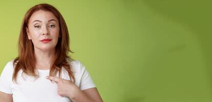 Proud mom pointing herself. Motivated confident redhead assertive middle-aged woman indicating chest volunteering bragging own abilities achievements look camera self-assured green background photo