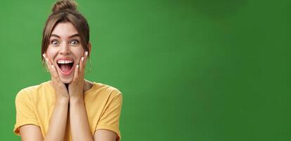 Portrait of happy delighted and surprised young feminine girl in yellow t-shirt pressing hands to cheeks from amazement and joy smiling broadly reacting to astonishing news over green background photo