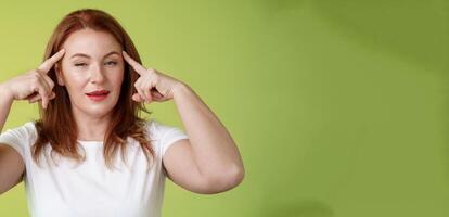 Smart choice. Intrigued thoughtful good-looking assertive redhead creative middle-aged woman touch temples squinting pondering info thinking use mental power read minds stand green background photo