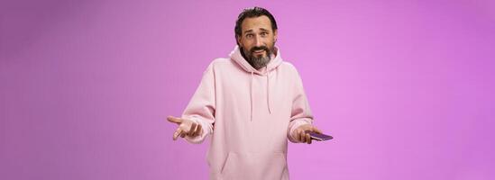 So what. Confused questioned adult bearded stylish guy in pink hoodie shrugging hands spread sideways dismay holding smartphone look clueless unaware camera not know how deal annoying phone calls photo