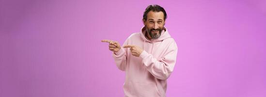 Sassy confident good-looking macho man 50s in pink hoodie smiling broadly inviting join pointing right showing interesting amusing place hang out welcoming take look, standing purple background photo