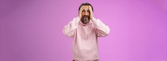 Adult upset bearded man despair standing troubled concerned touching face frowning grimacing sorrow unhappiness, losing bet, was robbed, standing shocked devastated purple background, panic photo