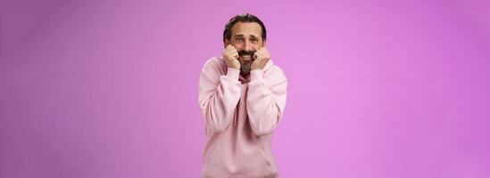 Scared insecure silly adult bearded man grey hair in pink hoodie press palms mouth bite fingers clench teeth shocked frightened widen eyes terrified standing stupor horrified, purple background photo