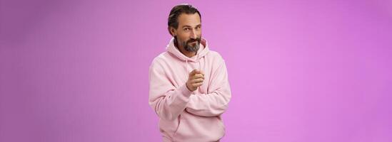 Confident handsome charismatic adult bearded man in stylish pink hoodie inviting you join company smiling self-assured indicating picking make decision chosing us, purple background photo