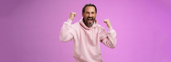 Cheerful supportive manly mature adult bearded guy fan yelling raising clenched fists triumphing team scored goal celebrating standing pleased shouting achieving success, posing purple background photo