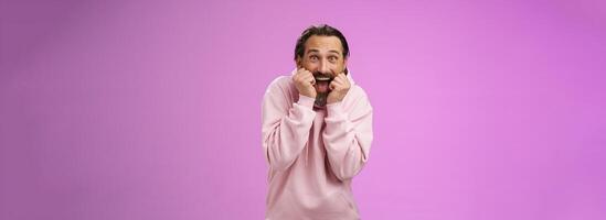 Amused thrilled gasping handsome happy bearded adult man in pink hoodie stooping screaming joyfully receive awesome incredible present standing speechless excited purple background photo