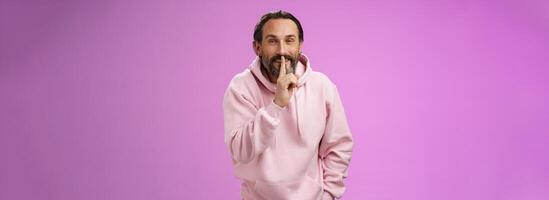 Lips sealed secret. Portrait alluring cheeky stylish adult bearded guy in pink hoodie asking keep quiet show shush shh gesture index finger pressed mouth smiling devious smug face, have idea photo