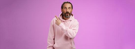 Amused handsome hipster mature 40s bearded caucasian man grey hair wearing trendy pink hoodie fold lips amazed wow pointing upper right corner widen eyes surprised check out awesome product photo