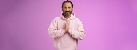 Peaceful attractive bearded mature mail model in trendy pink hoodie press palms together namaste praying gesture smiling delighted relaxed bowing buddhism faith, appreciating help, look thankful photo