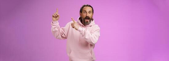 Impressed shocked exctied cool hipster mature 50s man bearded grey hair in pink hoodie retelling incredible story pointing up index fingers widen eyes surprised thrilled awesome advertisement photo