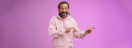 Charismatic happy good-looking mature 50s man wearing stylish hipster hoodie smiling excited pointing left index fingers impressed awesome interesting proposal standing pleased purple background photo