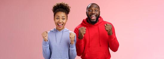 Lucky cheerful two african-american man woman yelling hooray celebrating triumphing huge success clenching fists joyfully accomplish mutual goal standing joyfully pink background victory gesture photo