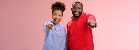 Joyful young attractive happy african-american couple vacation enjoying interesting tour standing impressed amazed pink background pointing index fingers camera awesome curious object photo