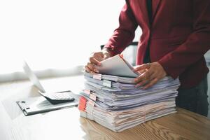 secretary is searching through pile of important documents on office table deliver them to manager for presentation in time at meeting. Concept of difficulty finding information from piles of document photo