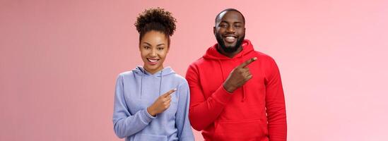 Charming carefree smiling african american two man woman grinning white teeth having fun fool around together pointing upper left corner showing couple project proudly, standing pink background photo