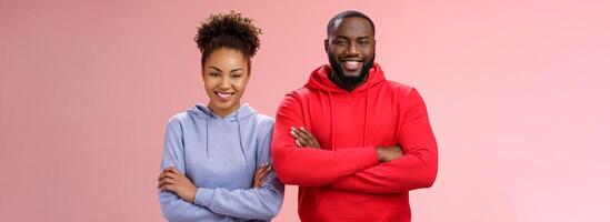 encantador contento profesional equipo dos africano americano hombre niña sonriente en general seguro de sí mismo propio habilidades cruzar brazos cofre sonriendo simpático imbatible trabajando juntos, en pie rosado antecedentes foto