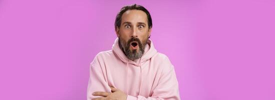 Shocked impressed gossiping caucasian bearded adult 40s man grey hair in pink hoodie gasping fascinated fold lips wow widen eyes astonished hearing interesting thrilling story, purple background photo