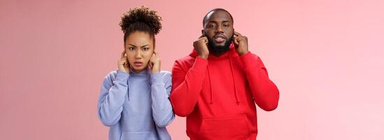 Annoyed tired couple african american woman man relationship exhausted each other close ears plug index fingers frowning look irritated hear terrible disturbing noise, standing pink background photo