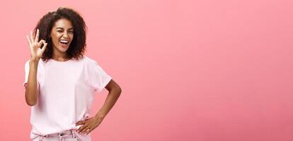 Girl got us covered. Charming charismatic and confident african american skillful woman in stylish outfit holding hand on waist showing okay gesture and winking assured at camera over pink wall photo