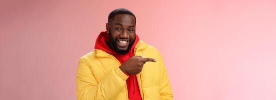 Charismatic carefree handsome black bearded guy in yellow jacket laughing friendly look camera chat pointing left show cool place hang out standing joyful pink background talking having fun photo