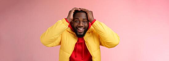 Waist-up surprised happy lucky young african-american bearded male in yellow jacket holding hands head smiling impressed joyfully receive awesome proposal dream come true, pink background photo