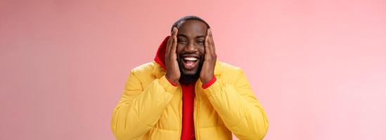 Happy charismatic playful african american bearded guy in yellow jacket red hoodie press palms cheeks smiling friendly laughing happily joking cannot believe receive awesome surprise, winning lottery photo