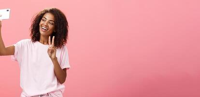 Stylish sociable good-looking dark-skinned female student with curly hairstyle pulling hand with smartphone near face taking selfie showing peace sign to device screen while smiling carefree photo