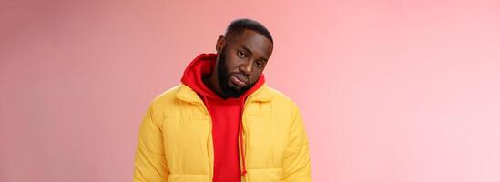 Unimpressed bored ignorant young african bearded man tilt head look scorn uninterest camera listen boring useless information, standing careless in yellow jacket red hoodie, indifferent photo