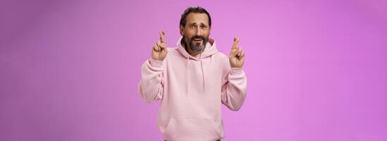 Nervous unsure hopeful handsome bearded 40s man in pink stylish hoodie cringing worried cross figers good luck make wish hope not gonna lose job supplicating praying dream come true photo