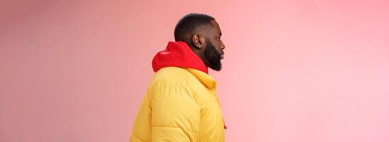 Profile studio shot bearded young 25s african guy in yellow jacket red hoodie look left normal unbothered relaxed expression standing queue order fastfood pink background, waiting take-away photo