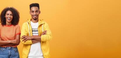 Portrait of surprised sarcastic african american woman with afro hairstyle standing with cute boyfriend crossing arms on chest laughing from scorn and fun over orange background photo