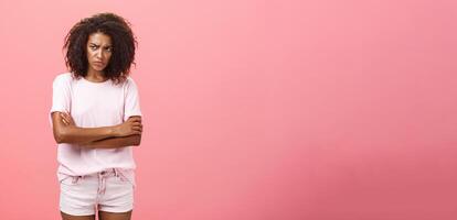 Portrait of offended gloomy sad african american female friend crossing arms on chest in protection gesture frowning looking from under forehead with insult looking envious and angry over pink wall photo