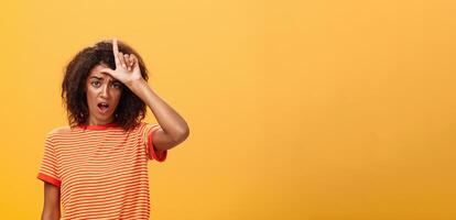 Girl thinks she loser. Portrait of gloomy bothered and displeased african american woman with afro hairstyle showing l word over forehead complaining feeling gloomy and unhappy over orange background photo