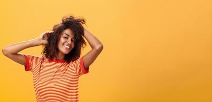 Waist-up shot of charming flirty feminine dark-skinned female in playful mood dancing playing with curly hair and smiling with delight and joy posing over orange background happy and carefree photo