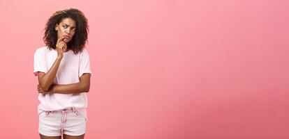 Suspicious displeased dark-skinned young curly-haired woman in trendy t-shirt and shorts saying hmm touching chin with finger frowning and staring left doubtful and unsure having concern photo