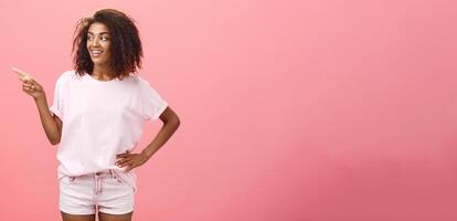 Girl checking out nice body of guy passing by. Charming confident dark-skinned female with curly hairstyle holding hand on hip looking and pointing left discussing cool copy space over pink wall photo