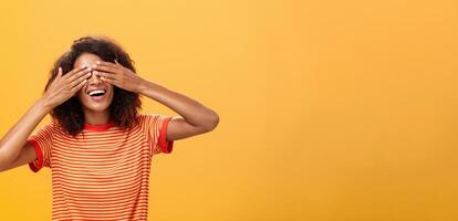 Closing my eyes and counting ten. Portrait of charming dreamy and happy funny african american curly-haired female in striped trendy t-shirt covering sight with palm and smiling waiting surprise photo