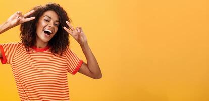 Nobody gonna spoil my perfect mood. Portrait of happy optimistic attractive dark-skinned female model with curly hairstyle tilting head smiling carefree showing peace or victory signs near face photo