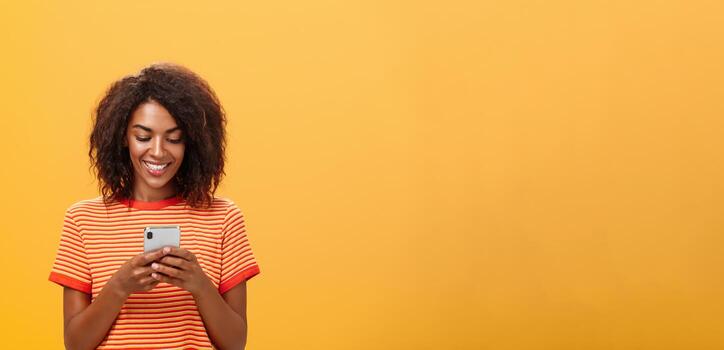 Typing answer to party invitation via smartphone message app holding device over chest smiling joyfully at gadget screen, texting or playing funny game, waste time in subway over orange wall photo