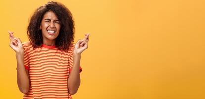 Waist-up shot of concerned worried woman eager take important job crossing fingers for good luck clenching teeth and closing eyes praying dreaming or making wish over orange background photo