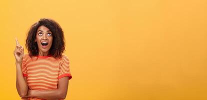 Waist-up shot of interested curious good-looking dark-skinned female in striped t-shirt talking asking question about curious star looking and pointing up with joy over orange background photo